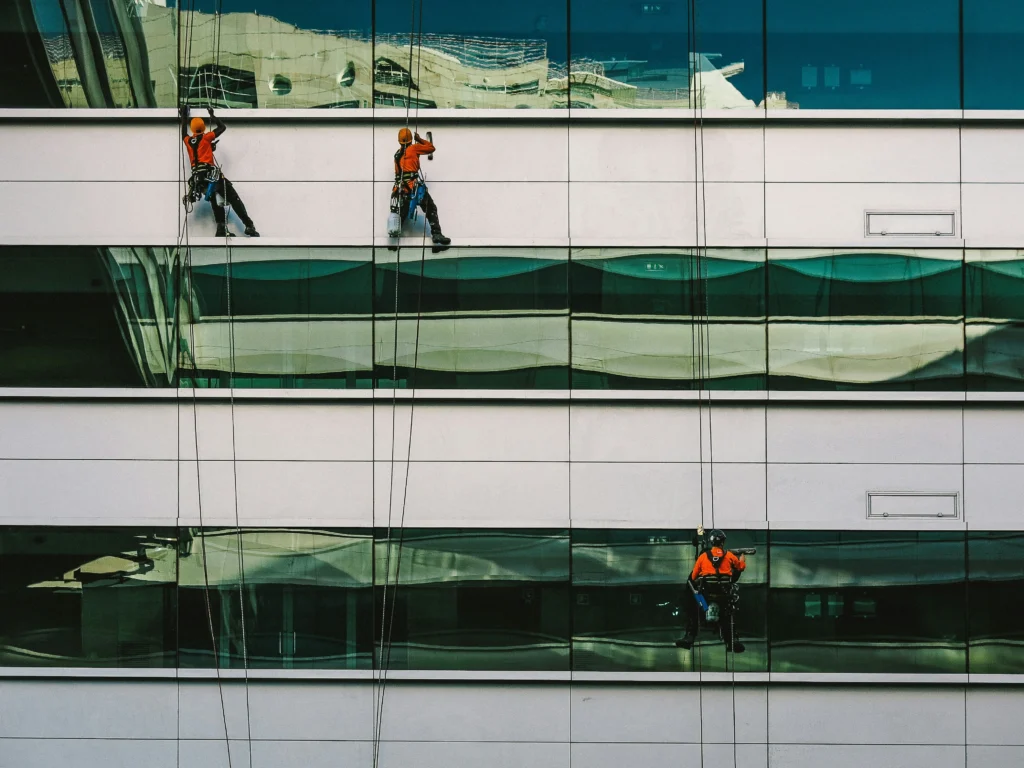 Three window cleaners wearing orange safety gear are suspended by ropes, cleaning the glass windows of a tall modern building.